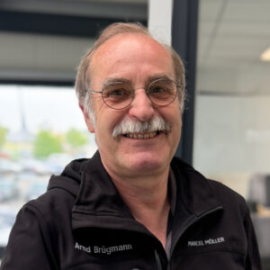 Arnd Brügmann _ Logistik Ein lächelnder älterer Herr mit Brille und Schnurrbart in schwarzer Arbeitskleidung mit Namensschildern der Firma Marcel Möller.