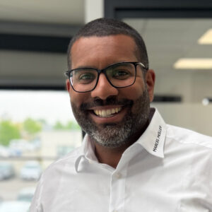 Marcel Möller_Geschäftsführer Ein lächelnder Mann mit Brille und Bart in weißem Hemd mit "Marcel Möller" Aufschrift, aufgenommen in einem Büroumfeld