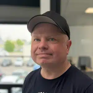 Marco Schwalba _ Fahrzeugaufbereiter Nahaufnahme eines lächelnden Mannes mit Baseballkappe und schwarzem T-Shirt vor unscharfem Hintergrund.