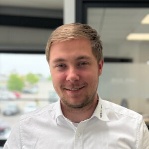 Matthis Källner_ Auszubildender Automobilkaufmann Junger Mann in weißem Hemd lächelt freundlich in die Kamera, im Hintergrund ein Bürofenster mit Parkplatz.