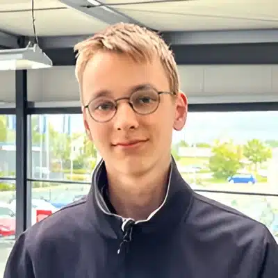 Lars Assmann Ein junger Mensch mit kurzen blonden Haaren und runder Brille in einer dunklen Jacke, fotografiert in einem hellen Raum mit Fenstern.