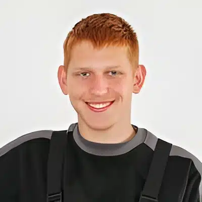 Felix Taubenheim Portrait einer lächelnden Person mit rötlichem Kurzhaarschnitt in schwarzem Oberteil vor hellem Hintergrund