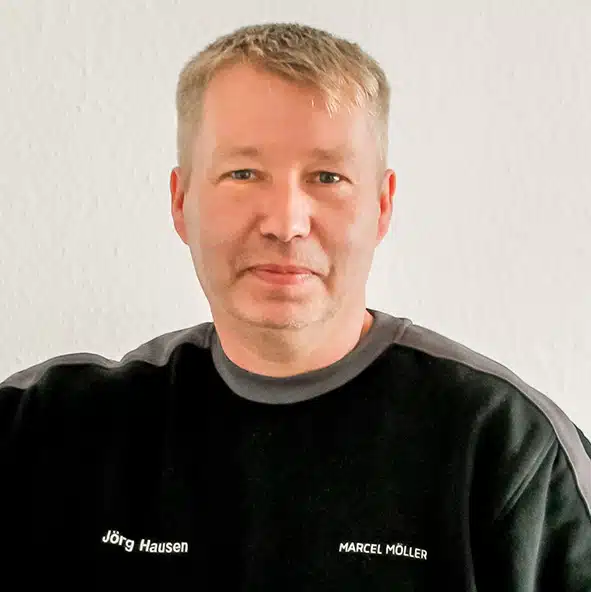 Jörg Hausen Ein Mann mittleren Alters mit kurzen blonden Haaren trägt ein schwarzes Shirt mit der Aufschrift "Jörg Hausen Marcel Müller" vor einer hellen Wand.