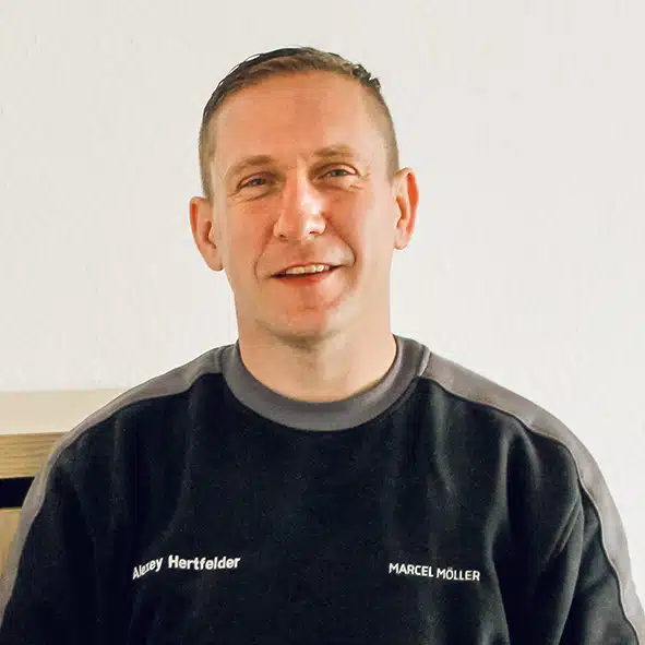Alex Hertfelder Portrait einer lächelnden Person in schwarzem Sweatshirt mit der Aufschrift "Away Hertfelder" und "Marcel Möller" vor hellem Hintergrund.