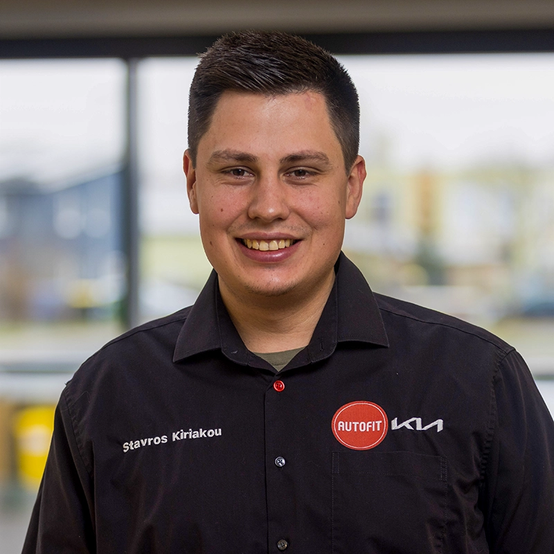 Stavros Kiriakou Porträtaufnahme eines jungen Automechanikers in schwarzem Arbeitshemd mit Autofit-KIA Logo und freundlichem Lächeln.