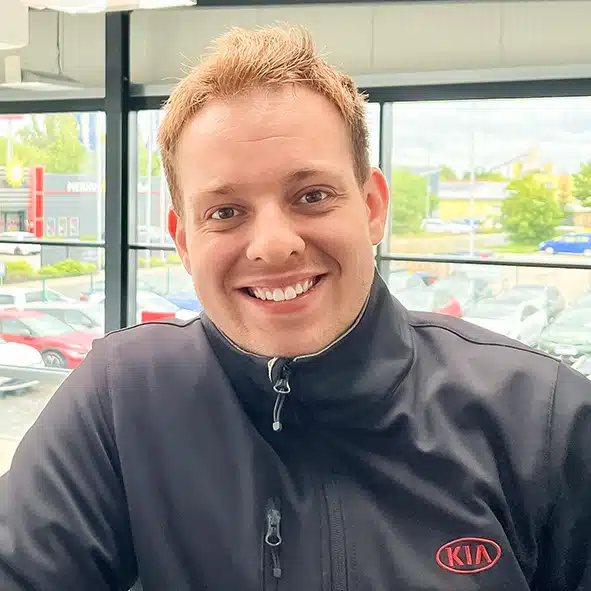 Lächelnder Mitarbeiter in schwarzer KIA-Jacke vor Autohausfenster mit Parkplatz im Hintergrund.