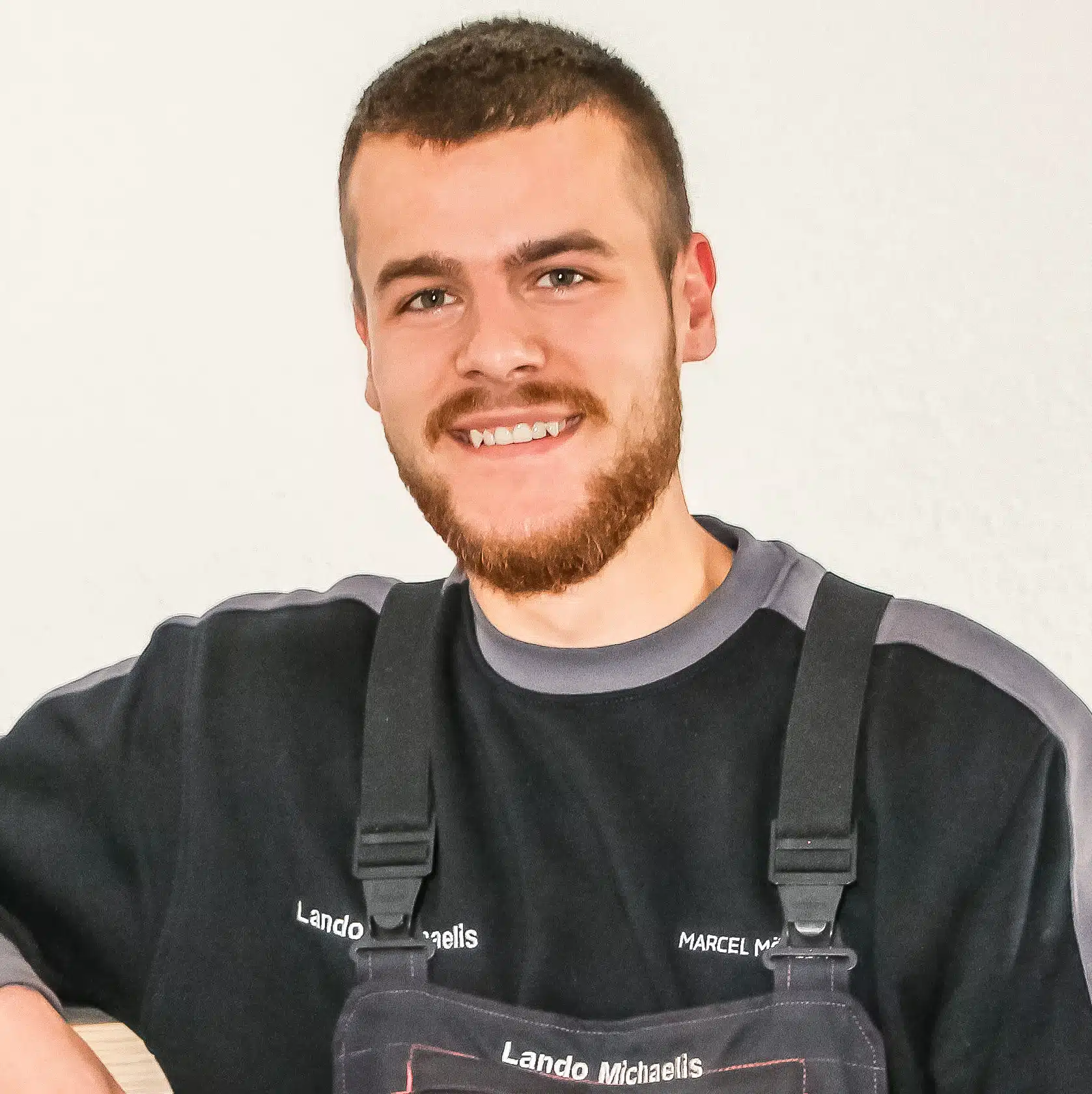 Lando Michaelis Junger Mann mit Bart in schwarzer Arbeitskleidung mit Trägern lächelt freundlich in die Kamera.
