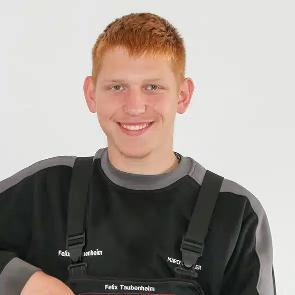 Freundlich lächelnder junger Handwerker in schwarzer Arbeitskleidung mit grauen Akzenten vor hellem Hintergrund.